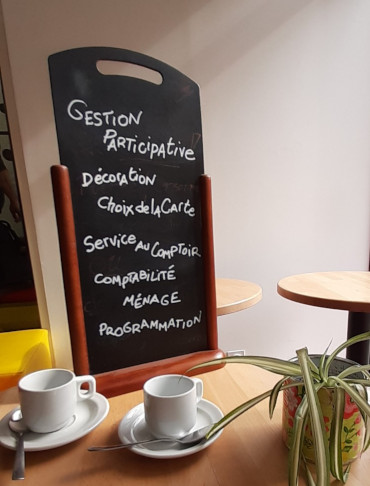 2 tasses à café devant une ardoise sur laquelle est écrit : "Gestion participative, décoration, choix de la carte, service au comptoir, comptabilité, ménage, programmation"